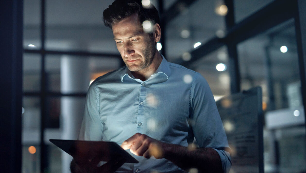 Ein Mann steht in einem dunklen Raum und hält ein Tablet in seinen Händen, dessen Displaylicht sein Gesicht anleuchtet.