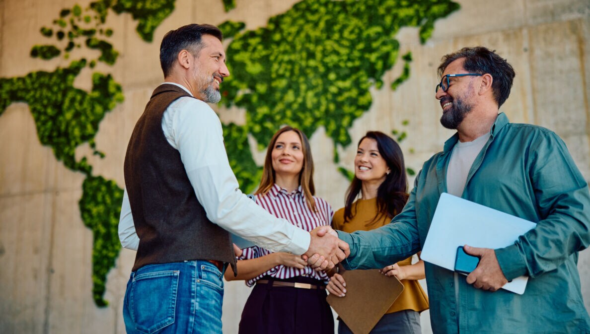 Zwei Männer mit Laptops im Arm geben sich lächelnd die Hand, daneben stehen zwei Frauen vor einer hohen Wand mit abstrakter, grüner Weltkarte.