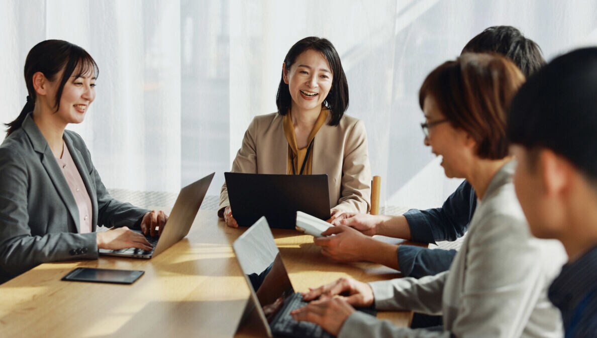 Eine Gruppe asiatischer Menschen sitzt lächelnd mit Laptops an einem Tisch.