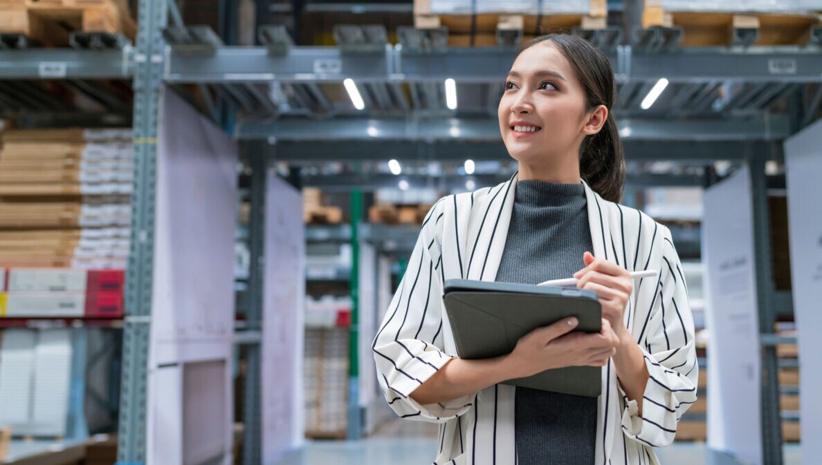 Eine lächelnde Frau steht mit Tablet und Stift in den Händen in einer Lagerhalle.