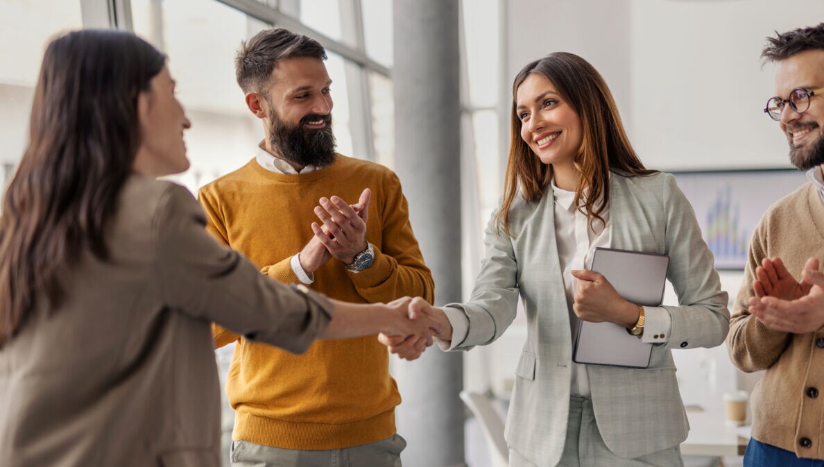 Vier Personen stehen beieinander, zwei Frauen geben sich lächelnd die Hand, während zwei Männer in die Hände klatschen.