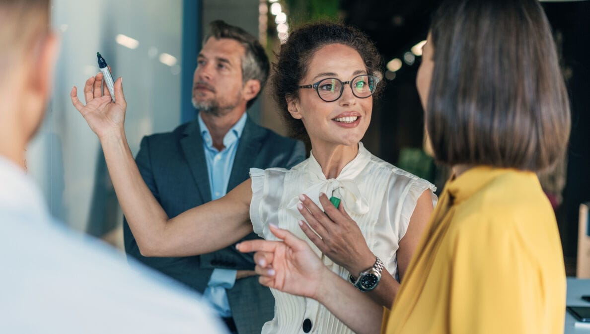 Vier Personen in Business-Kleidung stehen vor einer Wand und kommunizieren, eine Frau hält einen Stift in der Hand hoch.