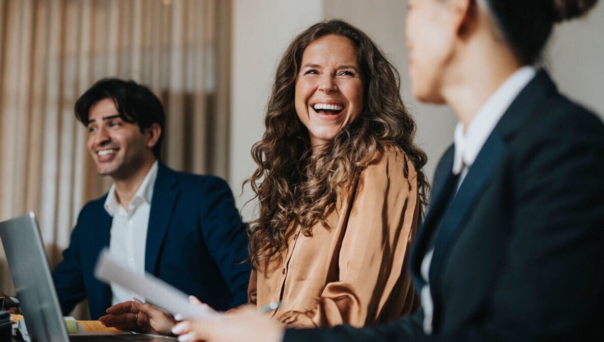 Eine lachende Frau sitzt vor einem Laptop an einem Tisch zwischen zwei weiteren Personen in Business-Kleidung während eines Meetings.