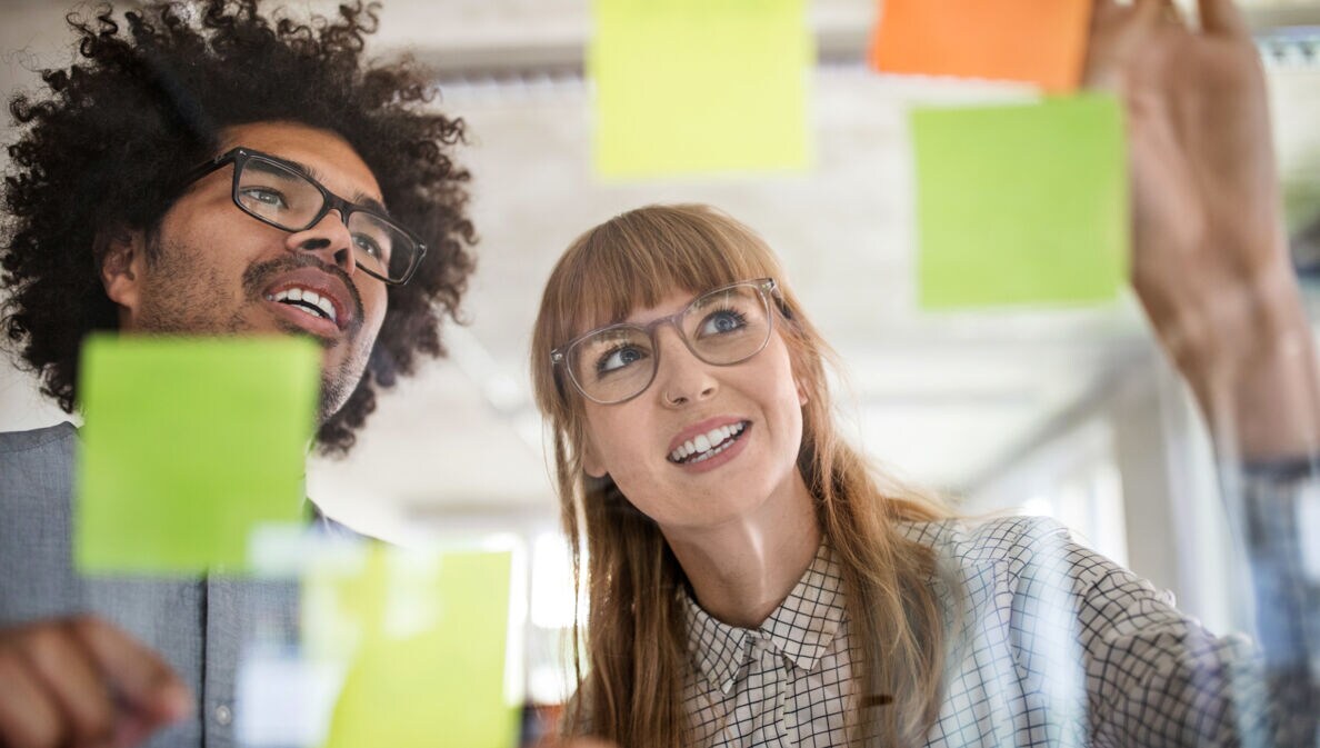 Nahaufnahme von Mann und Frau hinter einer Glasscheibe mit bunten Klebezetteln in einem Büro.
