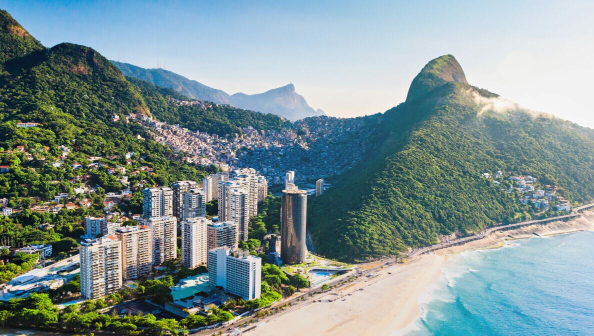 An einem Strand liegt eine Stadt mit Hochhäusern und riesigen Bergen.