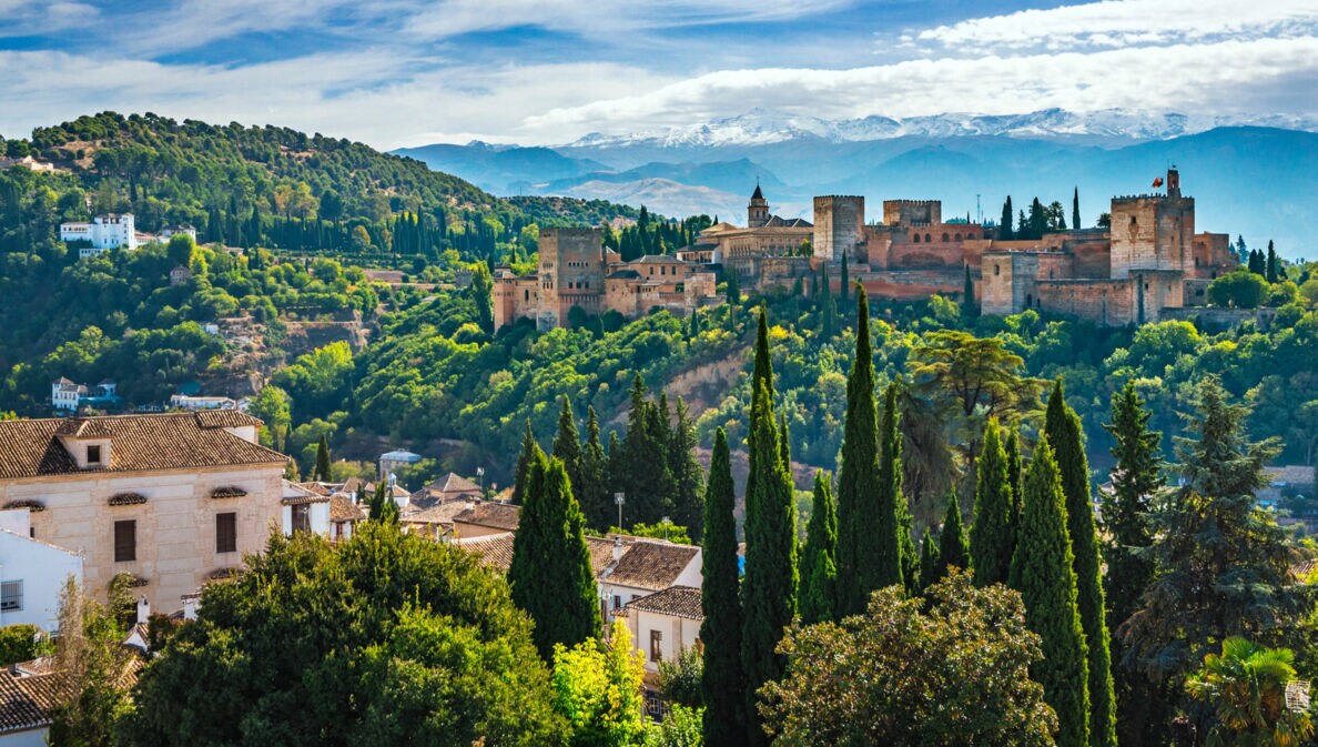Blick auf einzelne Häuser und die von Wald umgebene Alhambra, im Hintergrund Berge.