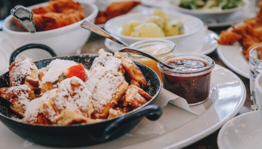 Ein schwarzer Topf mit Kaiserschmarrn auf einem weißen Teller, drumherum kleine Schälchen mit verschiedenen Leckereien.