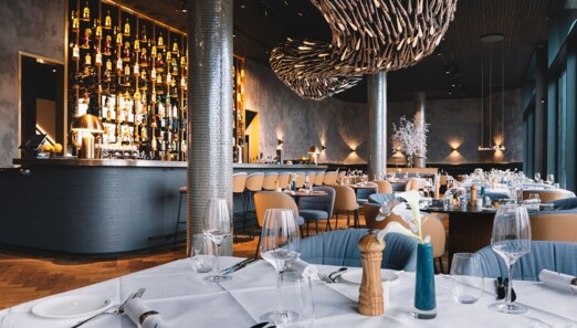 Stilvoller, modern eingerichteter Speisesaal mit Barbereich und eingedeckten Tischen unter einer abstrakten Deckenskulptur.