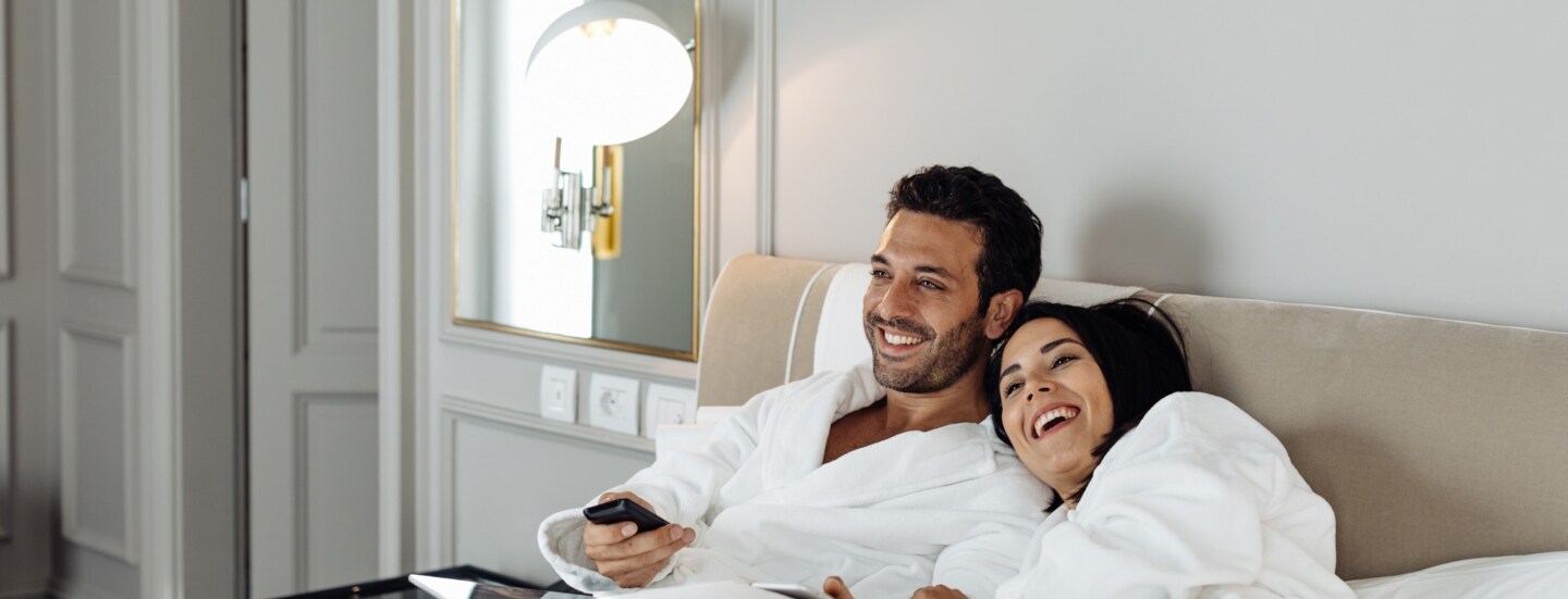 Ein fröhliches Paar in weißen Bademänteln entspannt im Bett einer edlen Hotelsuite