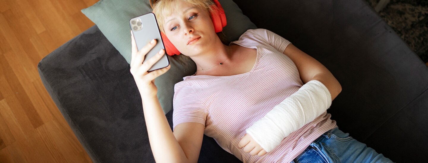 Aufsicht einer auf einem Sofa liegenden, jungen Frau, die mit Kopfhörern und einem eingegipsten Arm auf ihr Smartphone schaut.