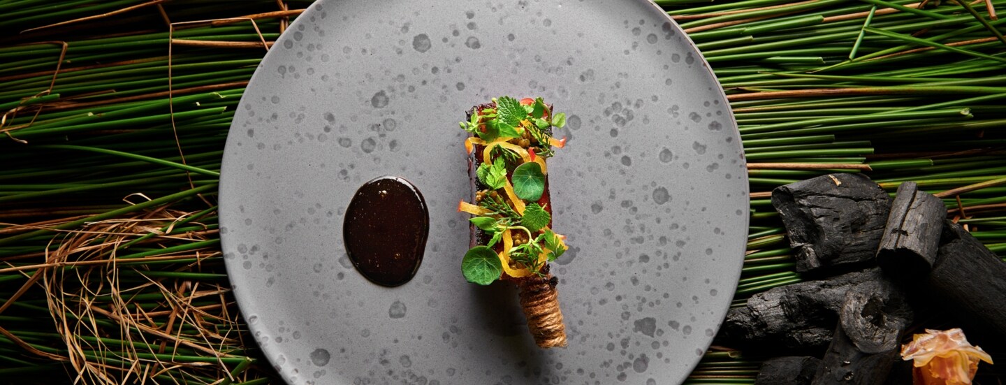 Aufsicht eines minimalistischen, vegetarischen Gerichtes auf einem grauen Teller, im Hintergrund grünes Schilfrohr.