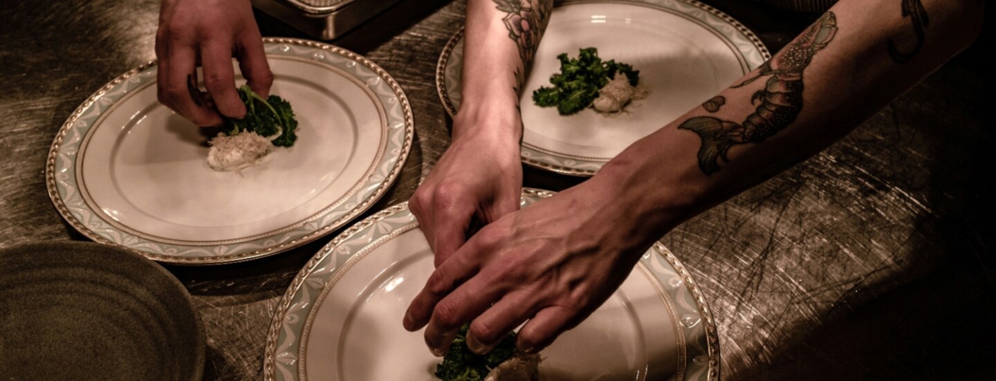 Nahaufnahme von Händen beim Anrichten von Speisen auf großen, weißen Tellern in einer Restaurantküche.