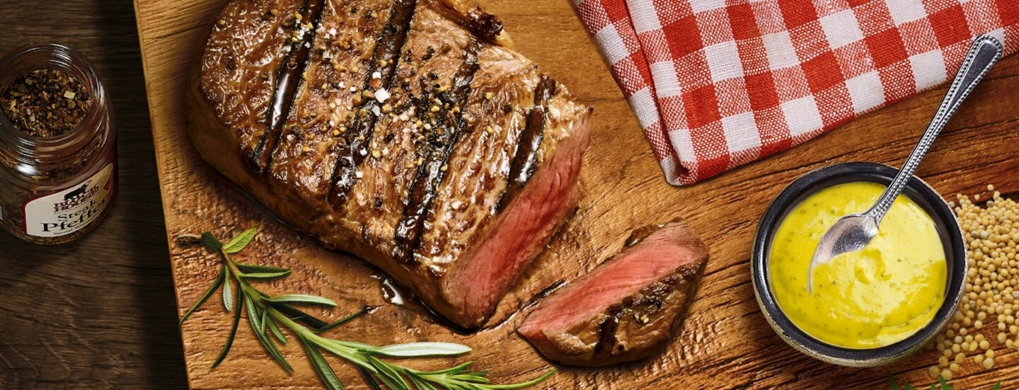 Aufsicht eines gebratenen, angeschnittenen Steaks auf einem Brett neben einer Schale mit Senf.