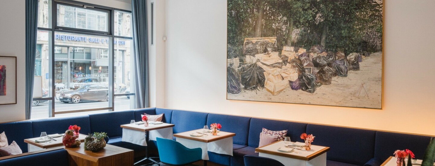 Innenraum eines hellen, modernen Restaurants mit blauen Sitzmöbeln an gedeckten Tischen und einem großen Gemälde an der Wand.