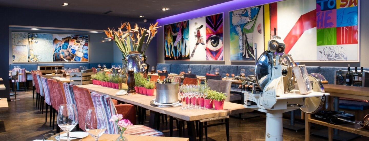 Restaurant mit einem langen, mit Pflanzen dekorierten Holztisch und einer Aufschnittmaschine mit Standfuß, umgeben von farbenfrohen Bildern an der Wand.