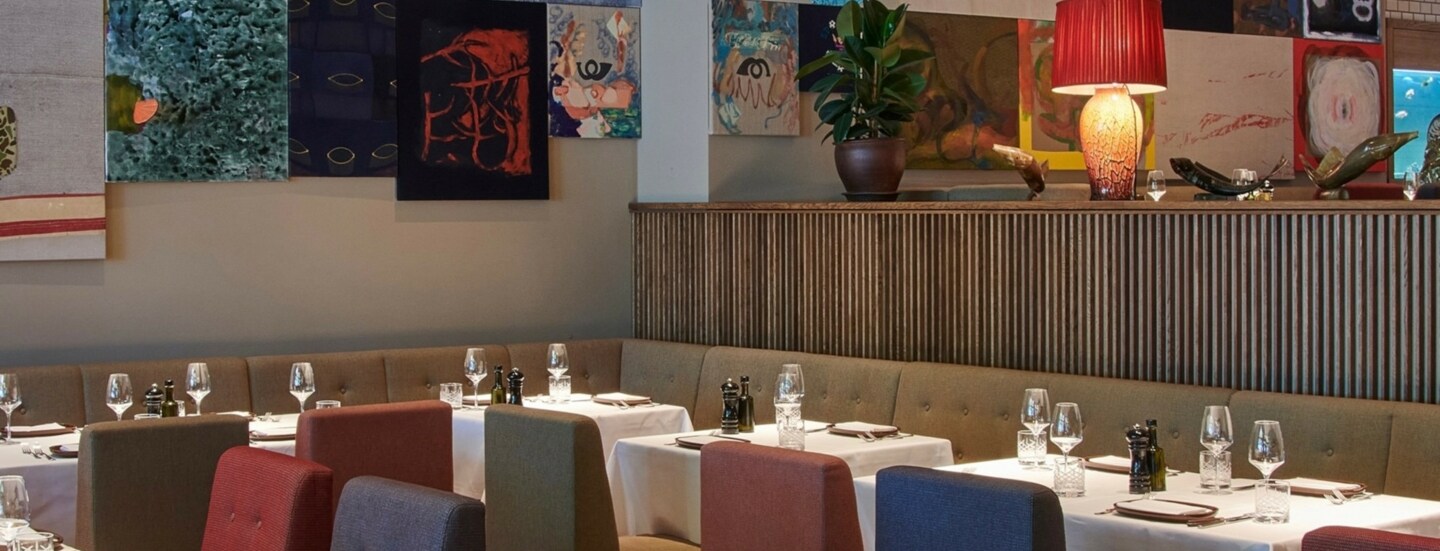 Restaurant mit Polsterstühlen an elegant gedeckten Tischen vor einer Wand mit farbenfrohen Gemälden.