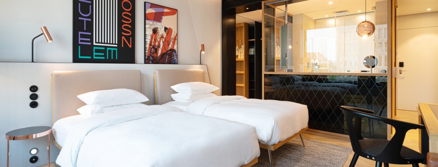 Elegantes Hotelzimmer im modernen Design mit zwei Betten und offenes Badezimmer hinter einer Glasscheibe.