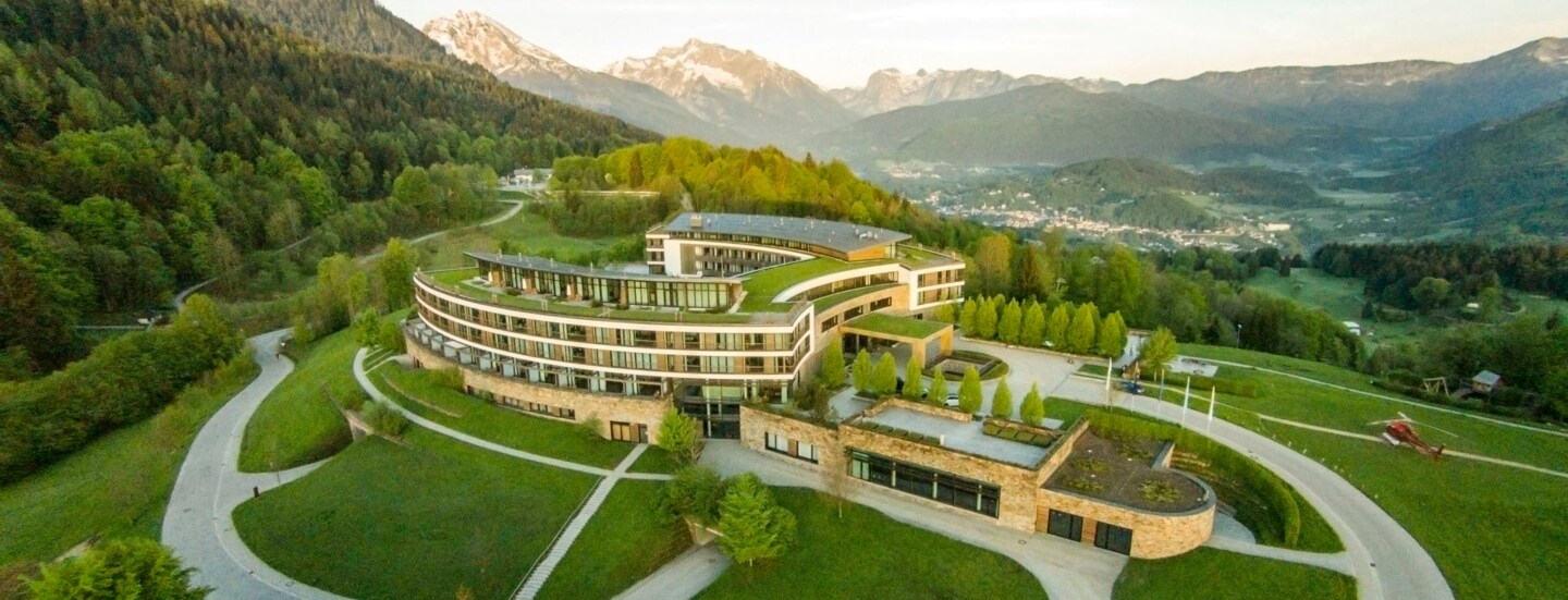 Luftaufnahme eines hufeisenförmigen Hotelkomplexes mit Rasenflächen inmitten einer hügeligen Waldlandschaft.