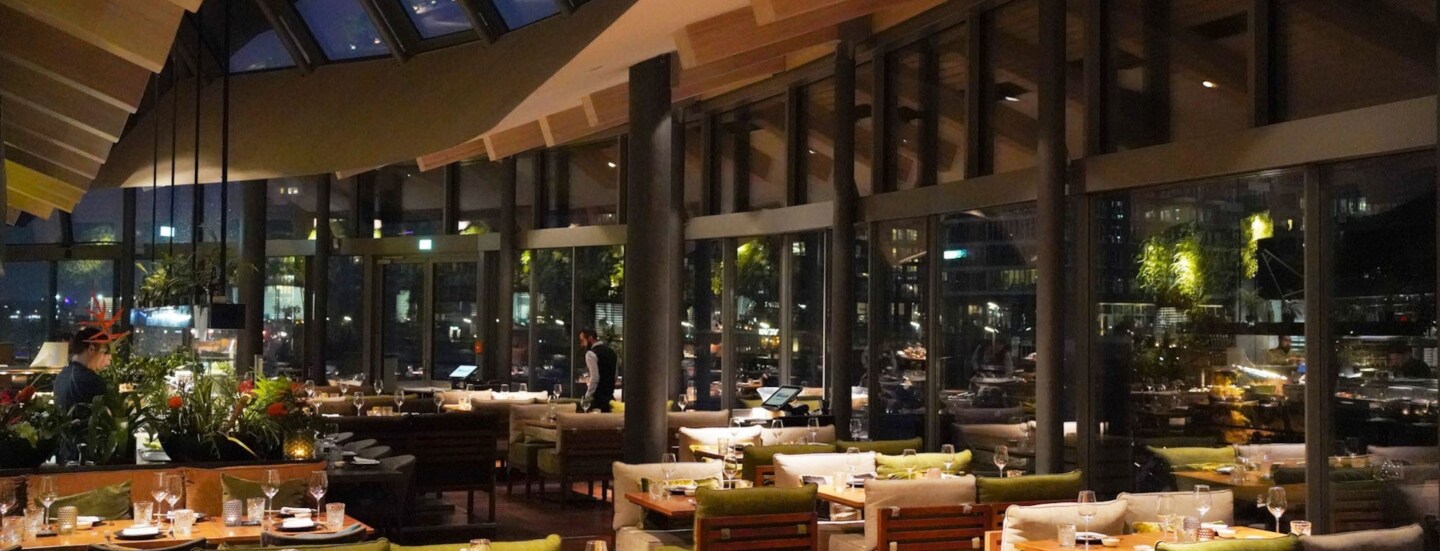 Speiseraum eines modernen Restaurants mit geschwungenem Dach und umlaufender Glasfassade bei Nacht.