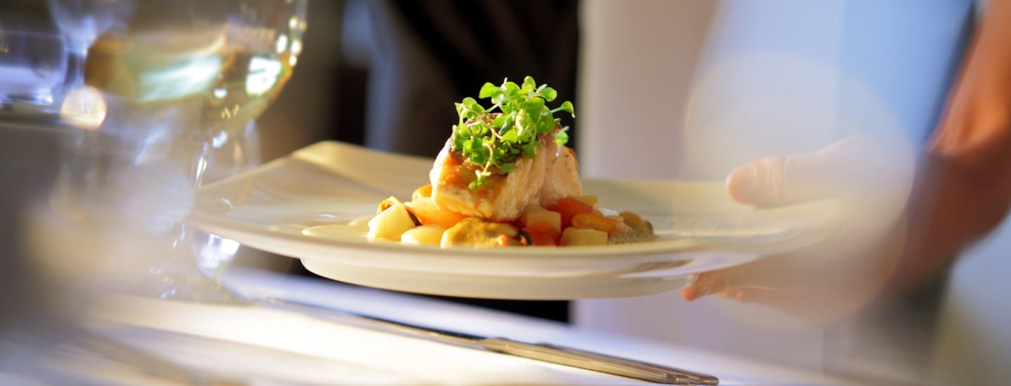 Nahaufnahme eines edel angerichteten Fischgerichtes, das auf einem weißen Teller an einem Tisch mit weißer Tischdecke serviert wird.