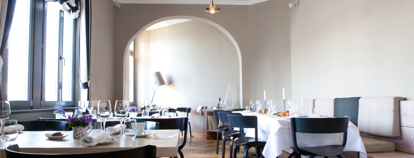 Heller Speisesaal eines edlen Restaurants mit schwarzen Holzstühlen an eingedeckten Tischen in einem klassischen Altbau mit hohen Decken und Holzparkett.