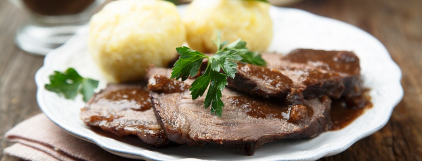 Sauerbraten mit brauner Soße an Klößen, angerichtet auf einem weißen Teller auf einem Holztisch.