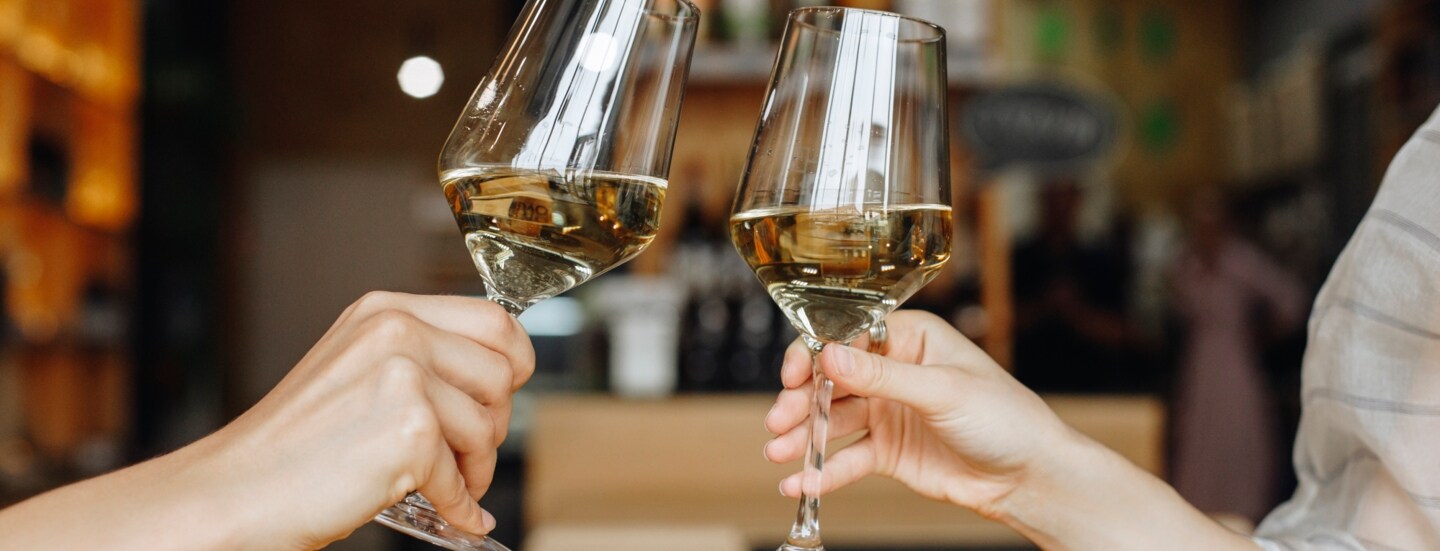 Nahaufnahme von Händen und Unterarmen von zwei Personen, die jeweils ein gefülltes Weinglas halten und anstoßen.