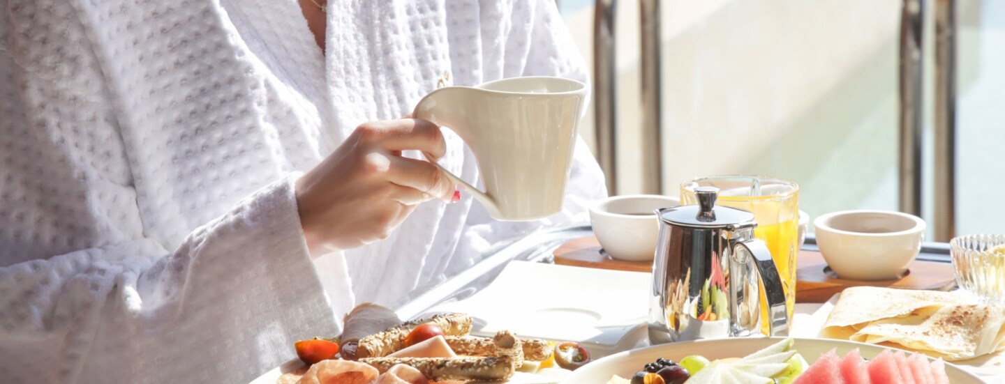 Eine Person sitzt an einem Tisch und hält eine Tasse in der Hand. Vor ihr steht ein Frühstückstablett mit Essen und Getränken.