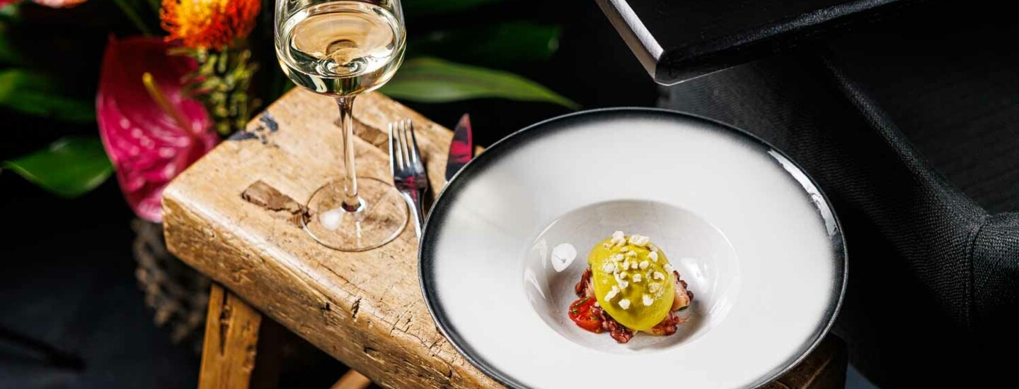 Ein weißer Teller mit einer kreativ angerichteten Vorspeise steht neben einem Glas Weißwein auf einem Holzschemel, daneben ein Blumenbouquet.
