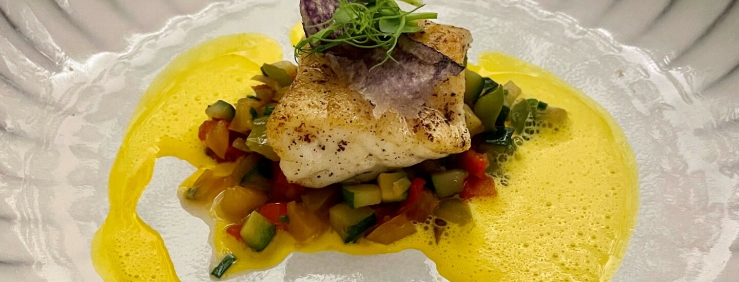 Nahaufnahme von einem Stück Steinbutt auf Gemüse, kreativ angerichtet mit gelber Soße auf einem weißen Teller.