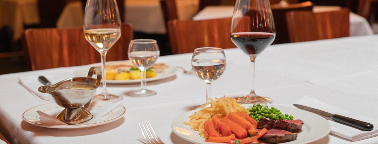 Zwei auf weißen Tellern angerichtete Speisen stehen neben Weingläsern auf einem Tisch mit weißer Stofftischdecke in einer Brasserie.