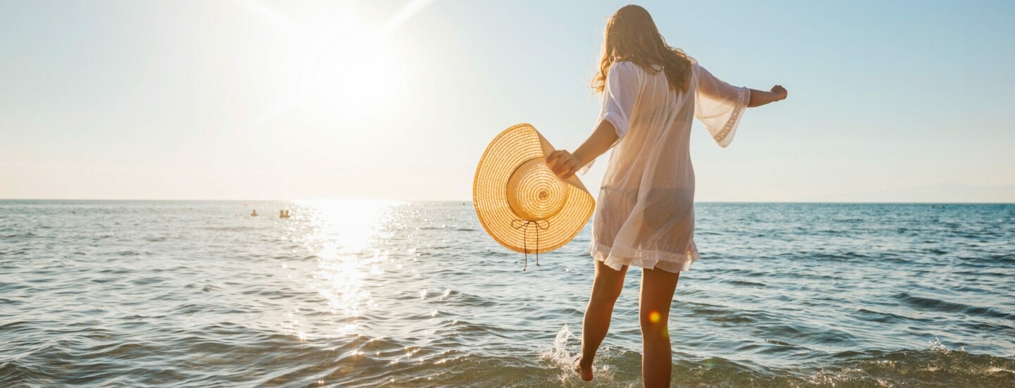 Eine junge Frau im weißen Sommerkleid und mit Strohhut in der Hand planscht mit ihrem Fuß im Wasser an einem Sandstrand am Meer bei Sonnenuntergang