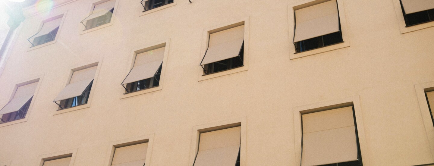 Hellbeige Hausfassade mit gleichfarbigen Markisen vor den Fenstern.