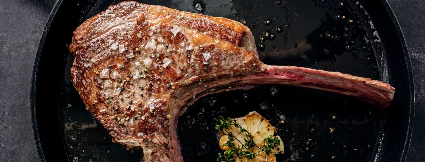 Ein scharf angebratenes Tomahawk-Steak in einer Pfanne. Daneben liegt eine aufgeschnittene Knoblauchknolle, die mit Thymian getoppt ist.