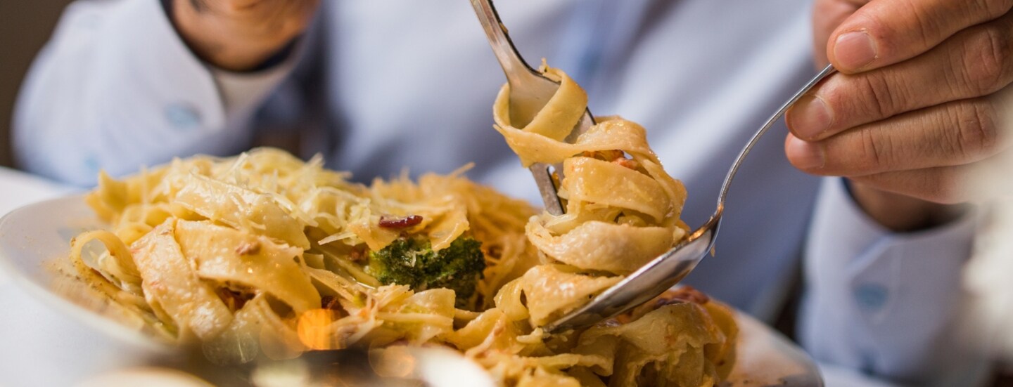 Nahaufnahme einer Person mit Hemd, die von einem weißen Teller mit einer Gabel auf einem Löffel Bandnudeln aufdreht.