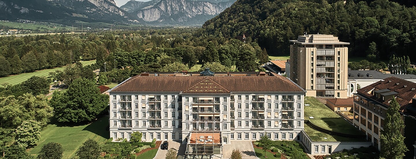 Luftansicht des Grand Resort Bad Ragaz in der Schweiz umgeben von grüner und waldiger Landschaft. Im Hintergrund Berge.