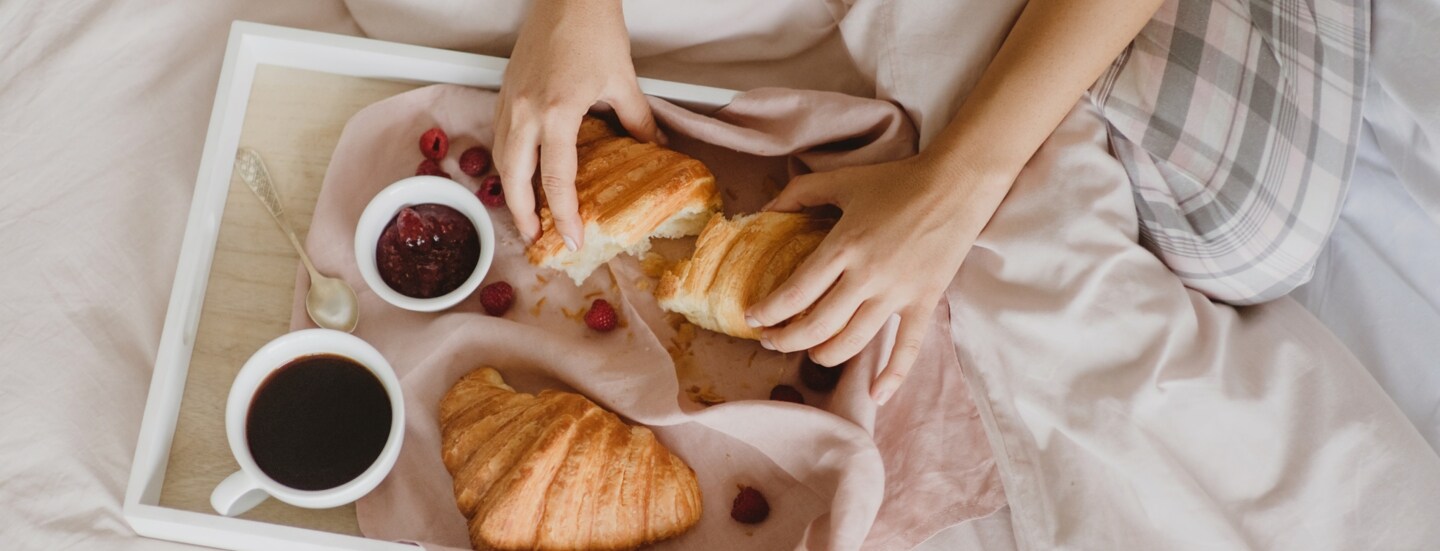 Auf einem Bett liegt ein Tablett mit Croissants, einer Tasse Kaffee, ein Schälchen Marmelade, einem Löffel und Früchten. Dahinter sitzt eine unkenntliche Person, die eines der Croissants auseinanderreißt.