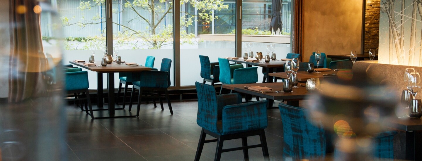 Blaue Polsterstühle an eingedeckten Tischen im Speisesaal eines Restaurants mit bodentiefen Fenstern.