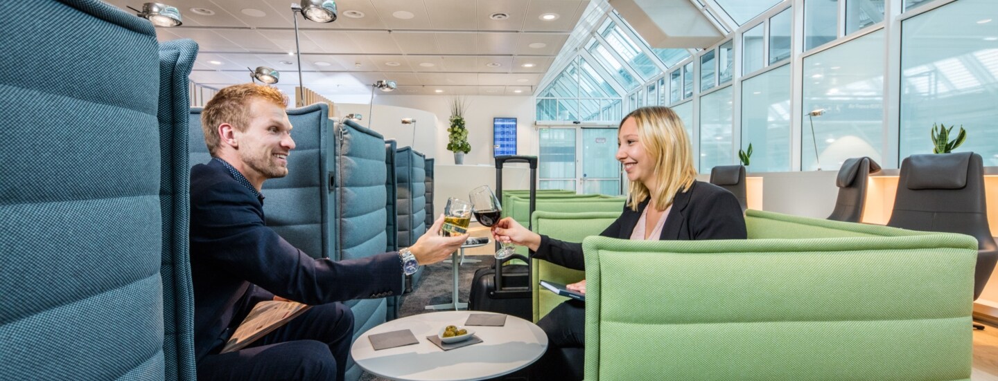 Ein Mann und eine Frau sitzen sich gegenüber auf Sesseln in einer Flughafenlounge und stoßen mit Weingläsern an.