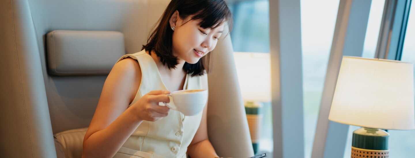 Eine Geschäftsfrau sitzt mit einer Tasse Kaffee in einem Sessel in einer Flughafenlounge und schaut auf ein Tablet in ihrer Hand.