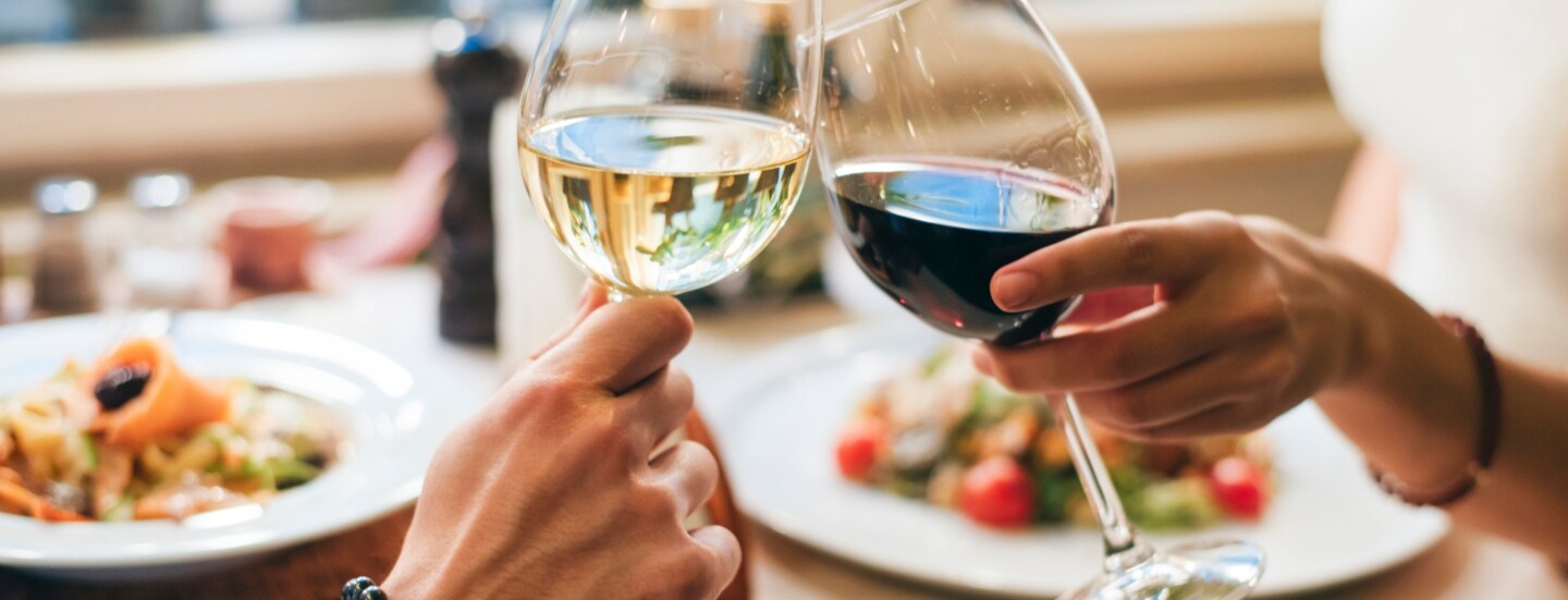 Zwei Personen, die jeweils ein Glas Wein halten und anstoßen. Auf dem Tisch vor ihnen stehen Teller mit Speisen.