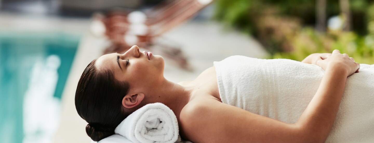 Eine Frau entspannt in einem weißen Handtuch auf einer Massageliege an einem Hotelpool.