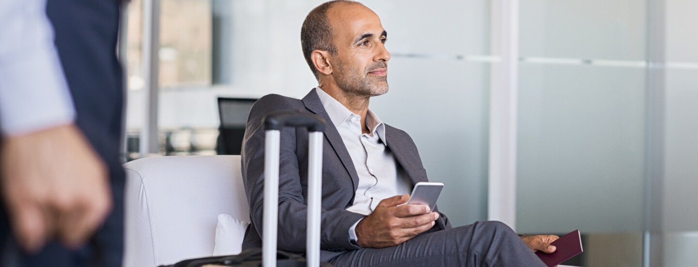 Ein Geschäftsmann sitzt in einem Sessel in einer Flughafenlounge, neben ihm steht ein Koffer.
