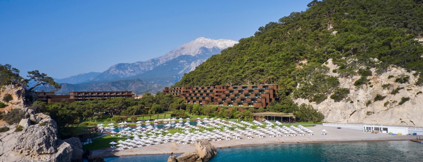 Ein Hotelresort in einer mediterranen Bucht mit Sandstrand, im Hintergrund eine Berglandschaft.