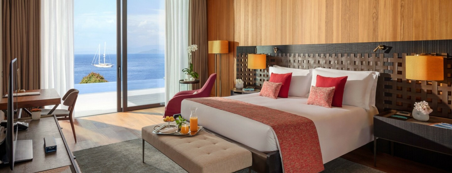 Modern eingerichtete Suite des Hotels Mandarin Oriental Bodrum mit einem großen Bett, einer Sitzbank, Arbeitstisch und raumhohen Fenstern mit Meeresblick.