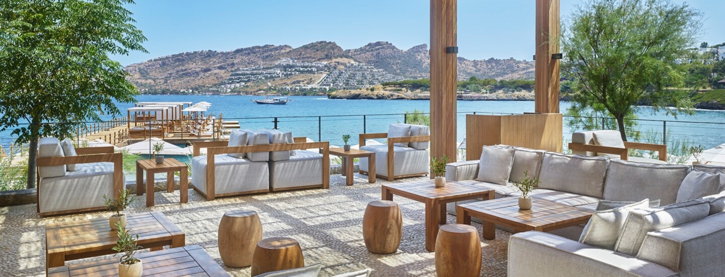 Loungebereich mit grauen Sesseln und Holztischen auf einer überdachten Terrasse an einer mediterranen Bucht.