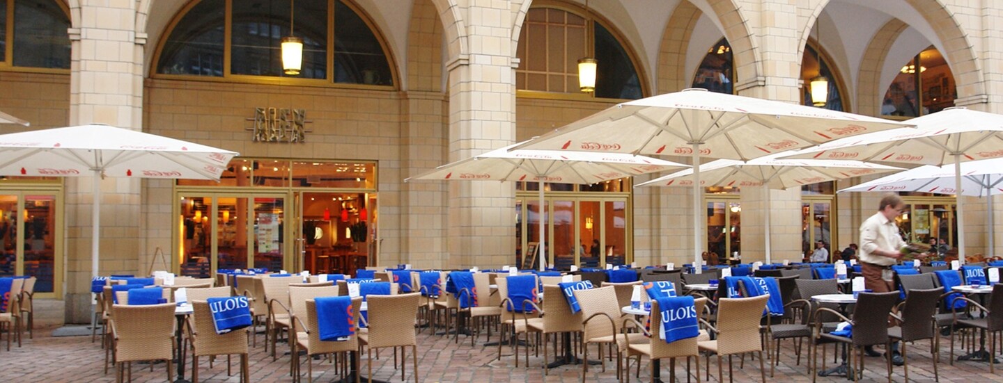 Außengastronomie auf einem Platz vor einem Gebäudekomplex mit Rundbögen.