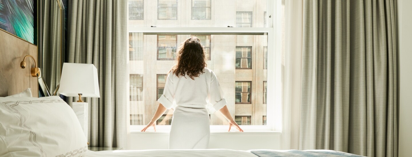 Rückansicht einer Frau im Bademantel, die am Fenster eines Hotelzimmers steht.