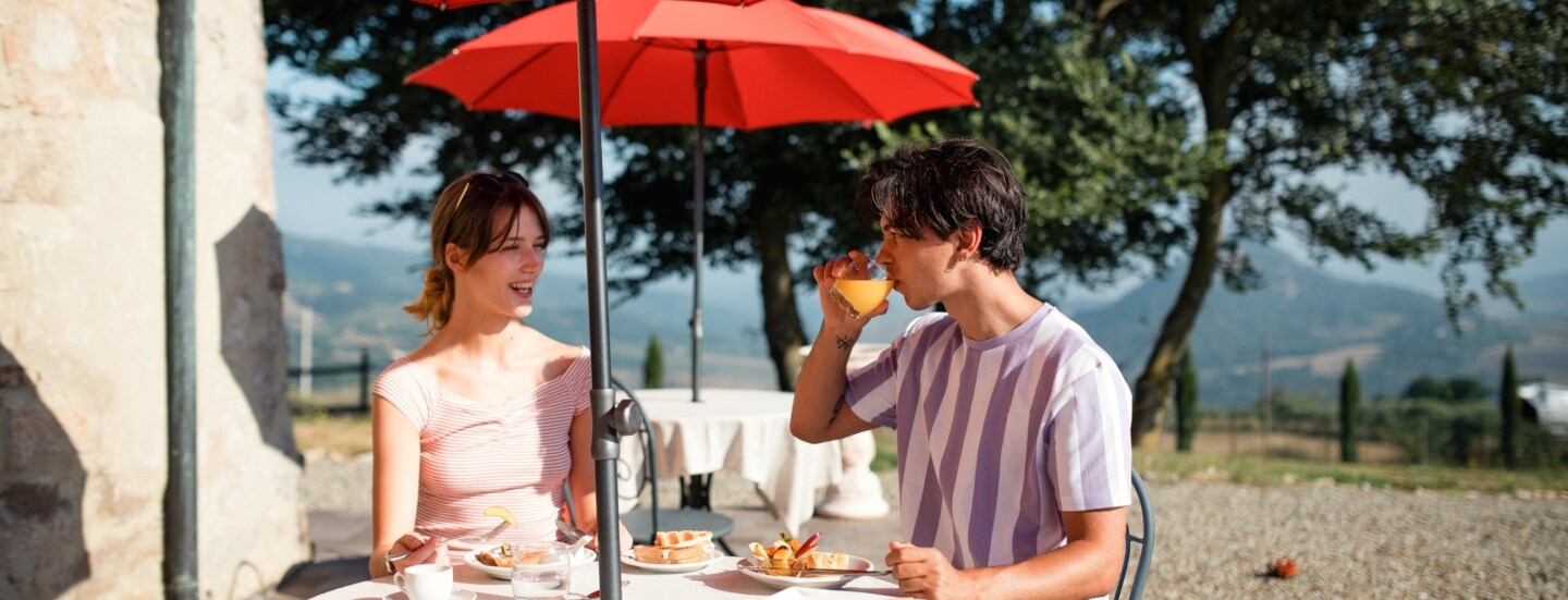Ein Paar frühstückt auf einer Terrasse an einem Tisch bei Sonnenschein, im Hintergrund eine Hügellandschaft.