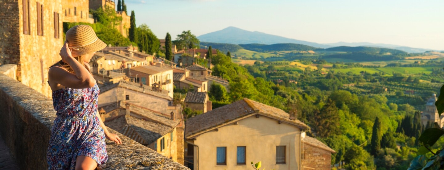 Eine Frau im Sommerkleid blickt von einer Mauer in einem italienischen Dorf hinunter auf eine grüne Hügellandschaft bei warmem Sonnenlicht.